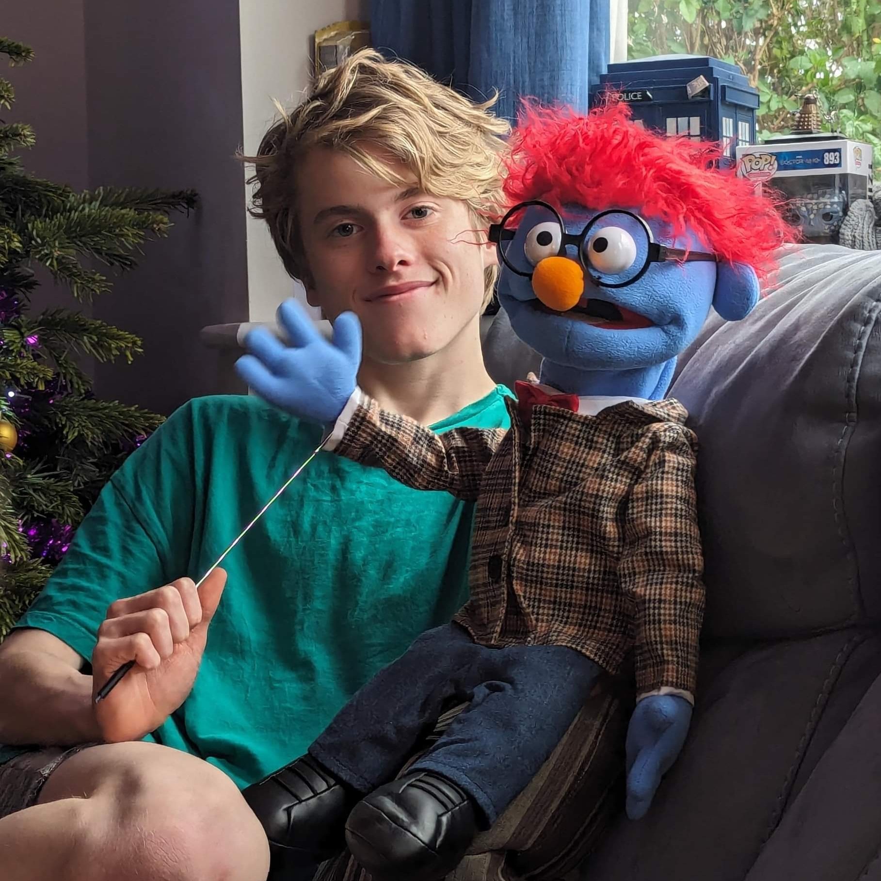 Young man with blue puppet with red hair sitting on a sofa and waving.