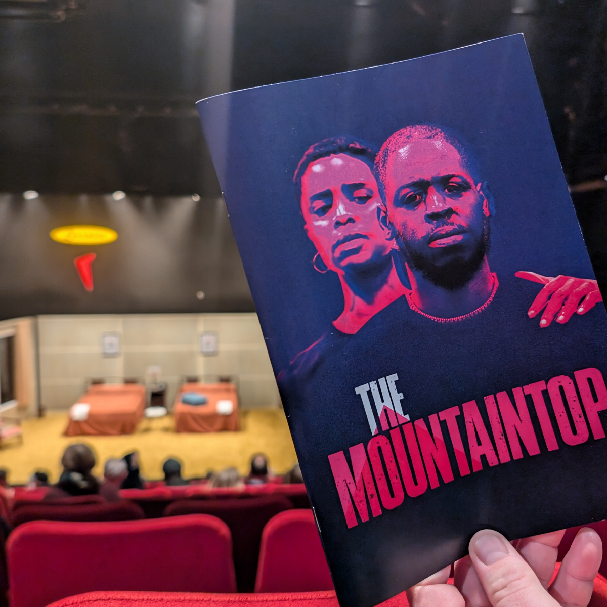 Programme of The Mountain Top with a woman standing behind a man. The programme is held in front of a stage set with two beds.