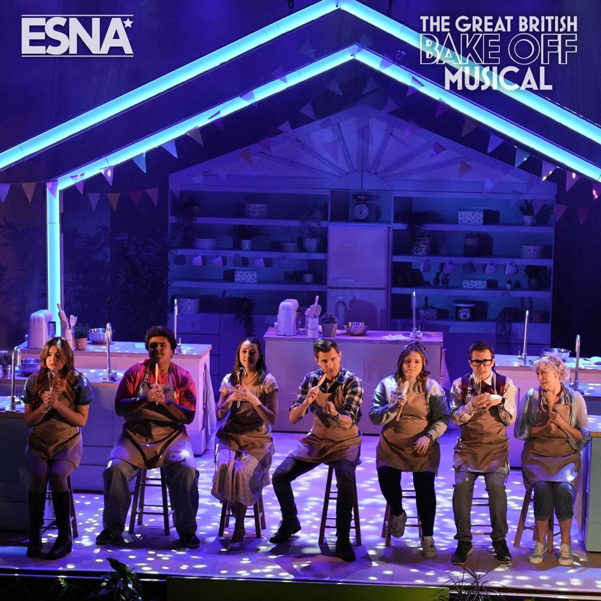 A group of seven people wearing aprons sitting on stools in a row. There is a stage set behind them which looks like an outline of a tent and some storage shelving. Thee is writing in the top right hand corner that says "The Great British Bae Off Musical" and in the top left corner "ESNA"