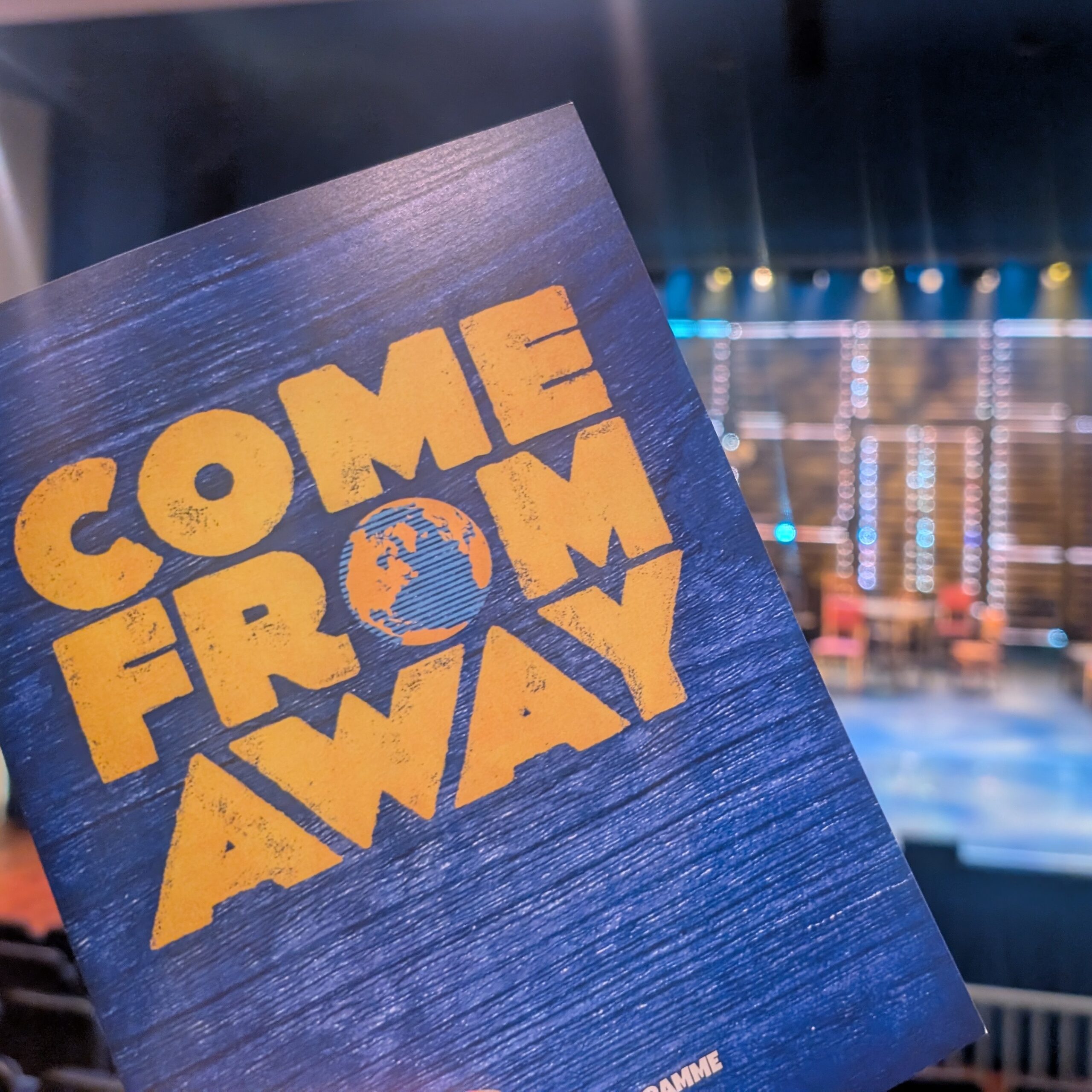 ComeFromAwayLaos There is a programme being held up in front of an empty theatre set. The words "Come From Away" are written in yellow across a blue background.